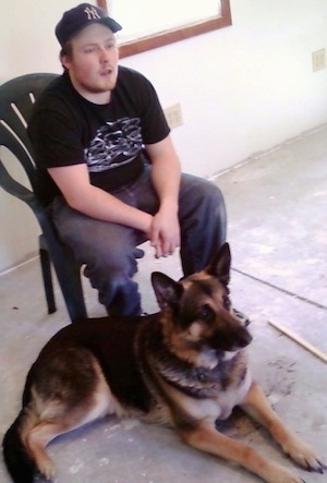 A black and tan German Shepherd is laying in front of a person sitting in a lawn chair in a house that looks to be under construction.